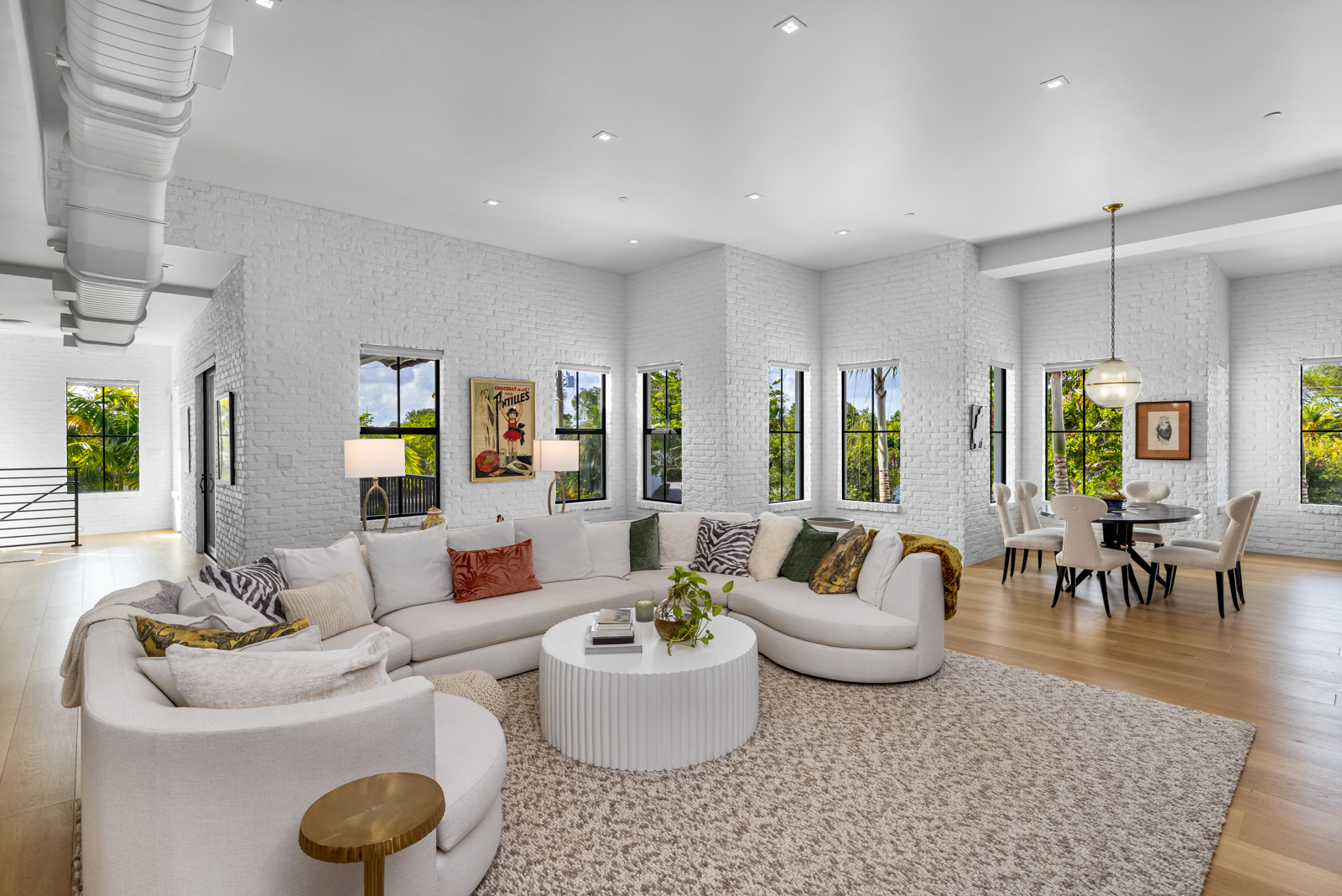 This is a bright and spacious living room featuring white brick walls, hardwood floors, and a large sectional sofa. The room is well-lit with natural light from multiple windows, and a round coffee table sits in the center of the room on a large area rug. The perspective is a wide shot, capturing the entire living space and adjacent dining area.