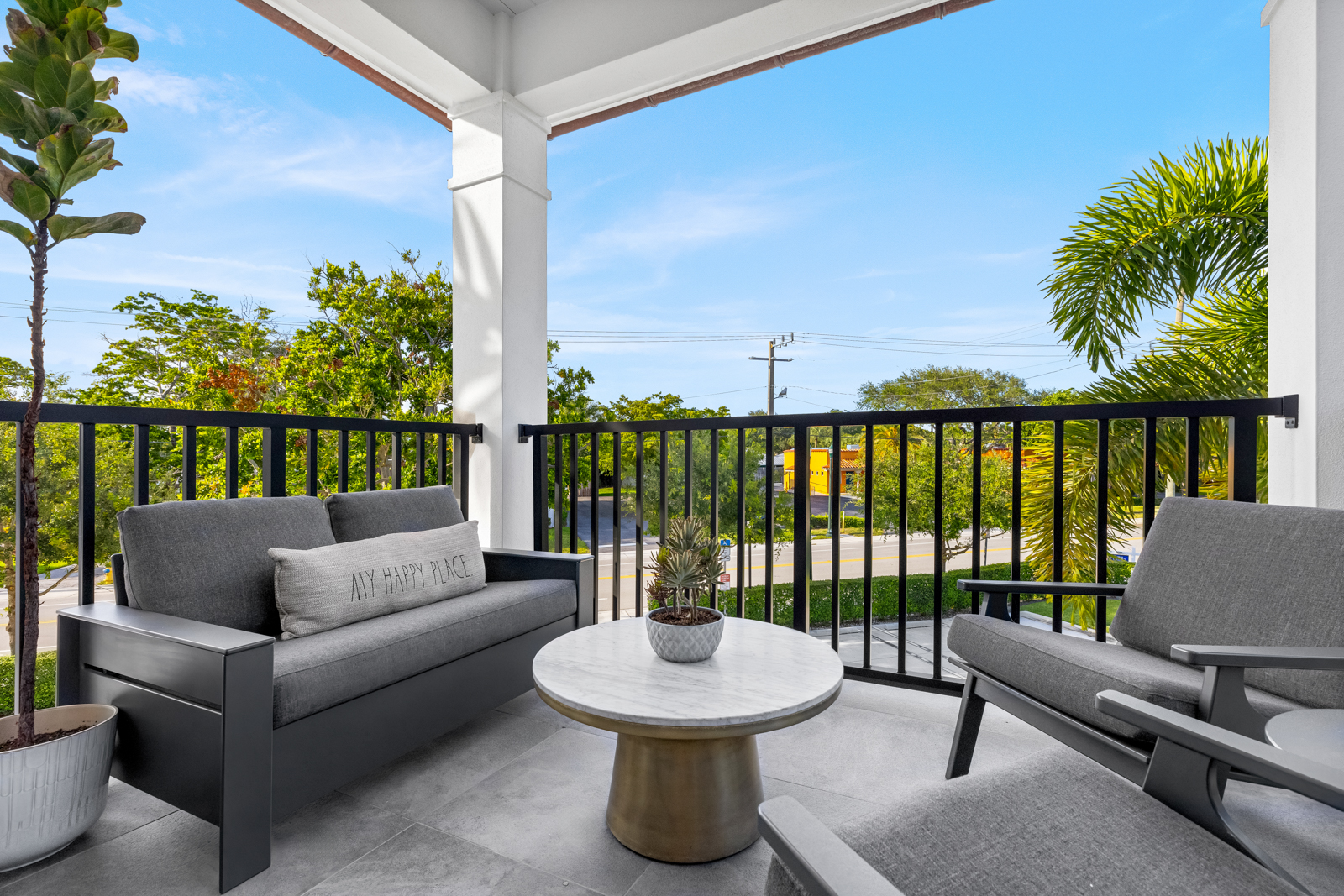 This image showcases a stylish balcony featuring modern outdoor furniture, including a gray sofa with a decorative pillow and two armchairs, arranged around a round marble-topped table with a potted plant. The balcony is enclosed by a black metal railing, offering a view of lush greenery and a clear blue sky, creating an inviting and relaxing outdoor living space.