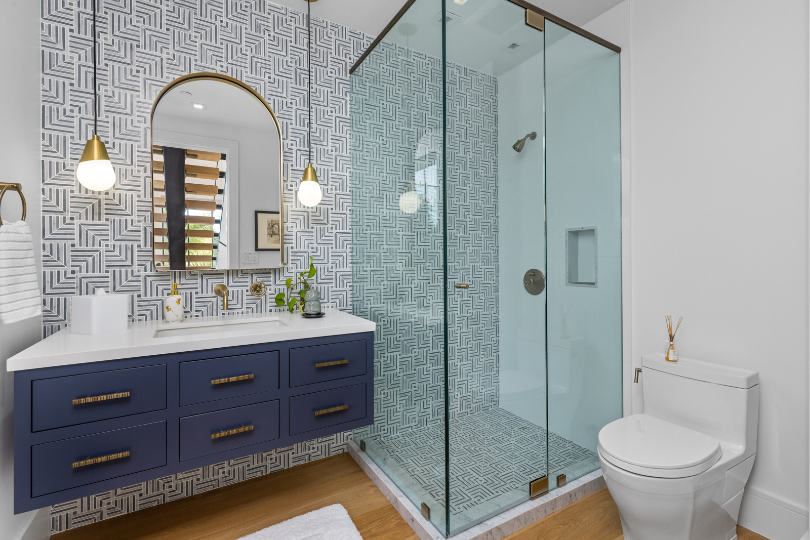 This is a stylish bathroom featuring a blue vanity with gold hardware, a white countertop, and a gold-framed arched mirror. The walls are adorned with a geometric patterned wallpaper, and the shower is enclosed with glass and features matching geometric tiles. A modern toilet sits to the right, completing the contemporary design.