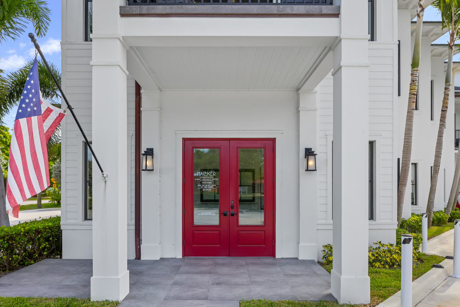 This image showcases the entryway of a building, featuring a pair of red double doors framed by white columns and a covered porch. An American flag is displayed to the left, adding a patriotic touch. The entryway is clean and well-maintained, suggesting a professional and welcoming atmosphere.