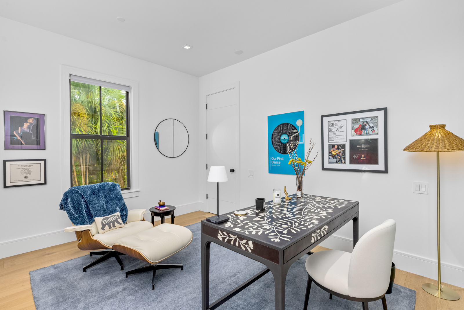 This is a well-lit home office featuring a modern desk with a unique patterned top, complemented by a white chair. The room includes a comfortable lounge chair with a throw blanket, a round mirror, and artwork on the walls, creating a stylish and functional workspace. The light hardwood floors and white walls contribute to a bright and airy atmosphere.