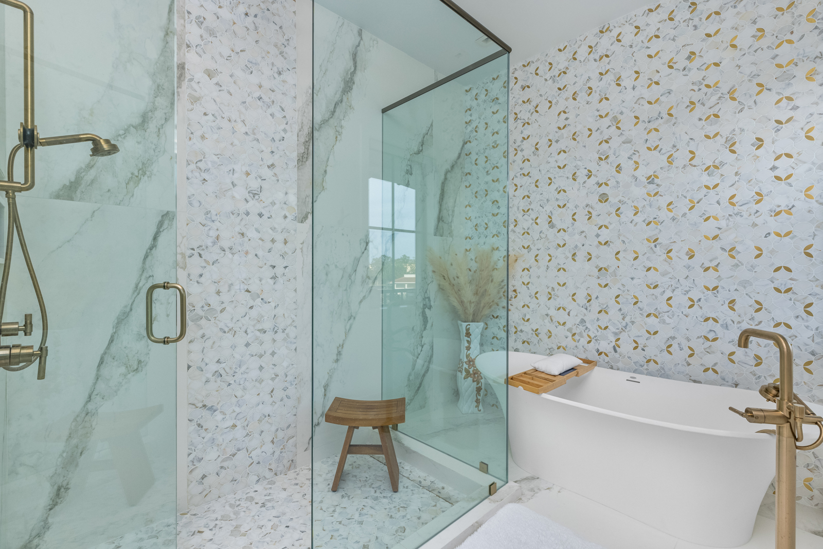 This is a luxurious primary bathroom featuring a glass-enclosed shower with bronze fixtures and a separate freestanding bathtub. The walls are adorned with decorative tile, creating a spa-like atmosphere. A wooden stool sits inside the shower, and a vase with pampas grass adds a touch of elegance.