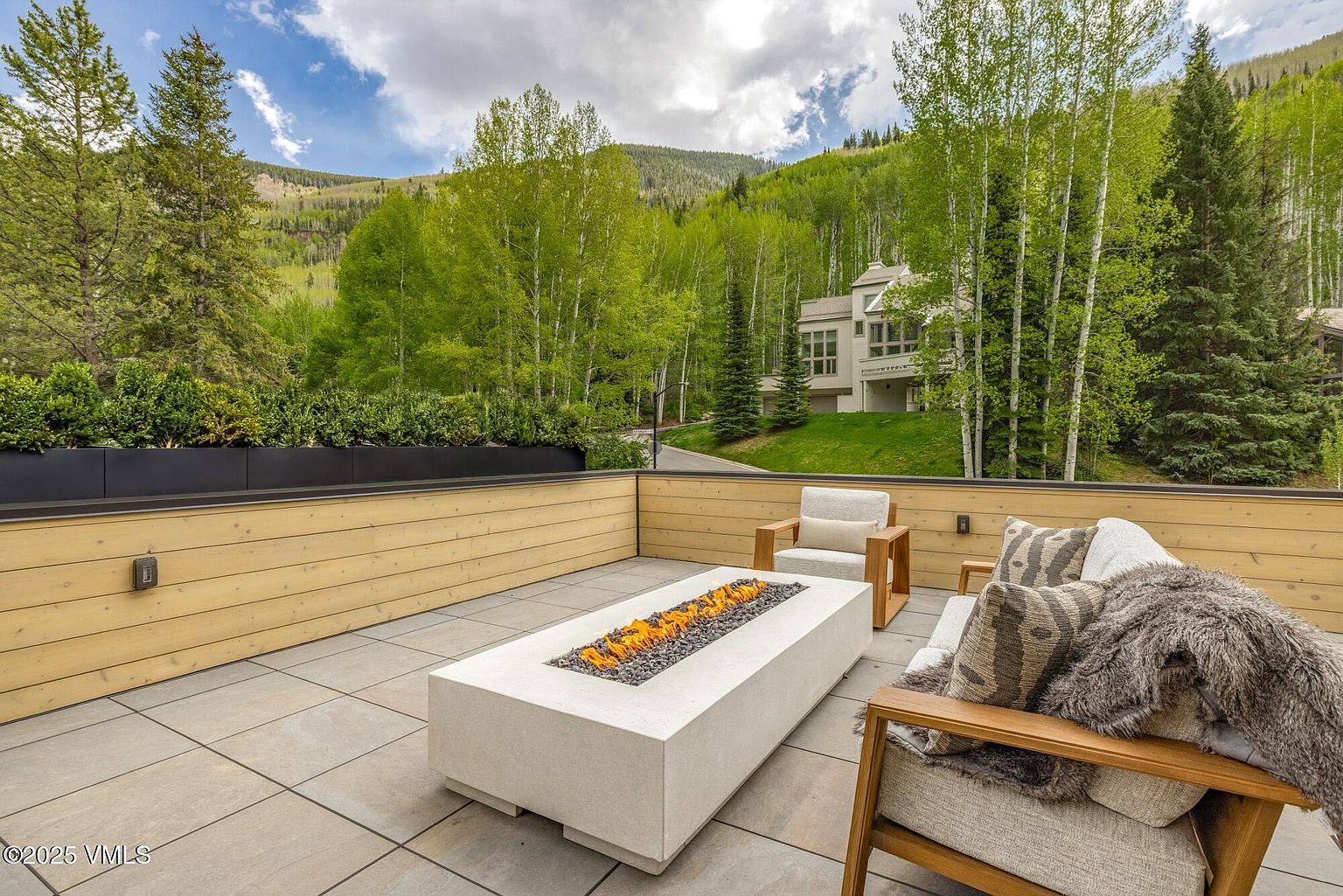 This elevated outdoor deck features a modern, rectangular fire pit as its centerpiece, surrounded by comfortable lounge seating with plush cushions and textured pillows. The space is enclosed by light-toned horizontal wood paneling and a planter box filled with lush greenery, offering a serene view of a forested mountain landscape. The perspective is from a slightly elevated vantage point, capturing the inviting atmosphere of this private, open-air retreat.