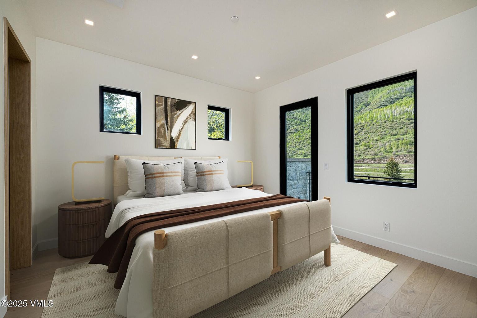 This modern bedroom features a neutral color palette with a light wood bed frame and a textured area rug. The room is illuminated by recessed lighting and natural light streaming through large windows that offer a scenic view of the surrounding hills. A minimalist aesthetic is maintained with clean white walls, simple bedside tables, and a piece of abstract wall art.