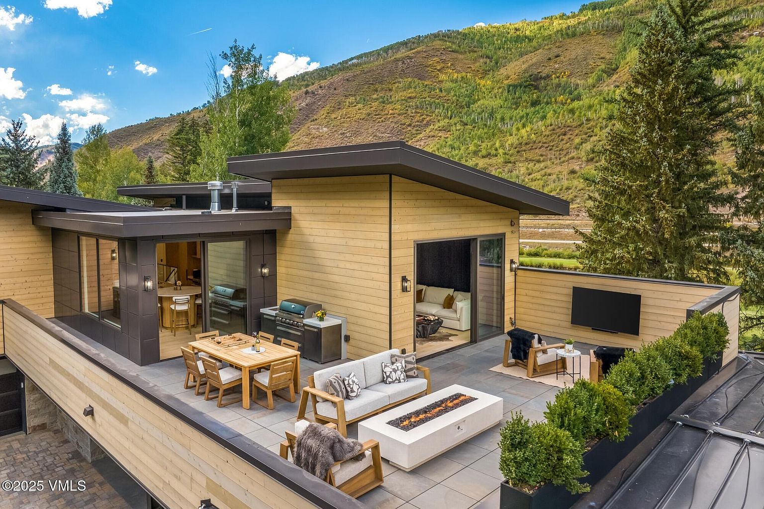 This elevated rooftop terrace offers a sophisticated outdoor living space with a modern aesthetic, featuring light wood siding and dark metal accents. The area is furnished with a dining set, a comfortable sofa, and lounge chairs arranged around a rectangular fire pit, all set against a backdrop of scenic mountain views. Large sliding glass doors provide seamless indoor-outdoor flow, while a wall-mounted television and integrated planters complete this luxurious entertainment space.