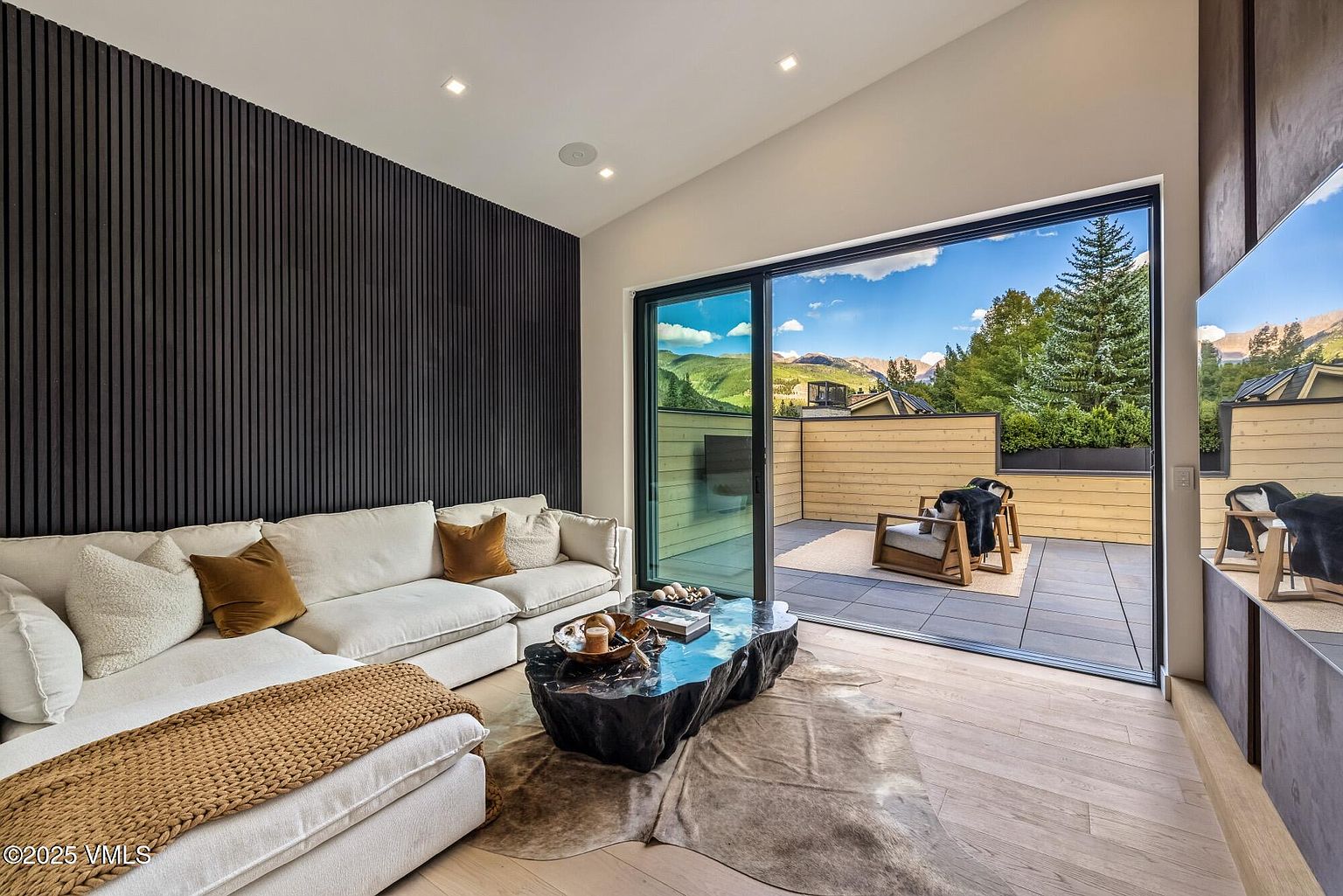 This contemporary living room features a striking dark wood-slat accent wall and a plush white sectional sofa adorned with textured throw pillows. A unique, organic-shaped black coffee table sits atop a cowhide rug, while large sliding glass doors open to a private outdoor terrace with mountain views. The space is bright and airy, blending modern interior design with a seamless transition to the exterior deck.