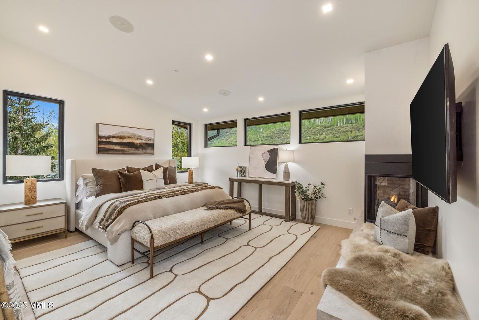 This primary bedroom features a neutral color palette with a large upholstered bed, layered bedding, and a textured bench at the foot. The room is accented by a fireplace with a dark surround, a wall-mounted television, and a console table beneath a piece of abstract art. Large windows provide natural light and views of the surrounding landscape, creating a serene and sophisticated atmosphere.