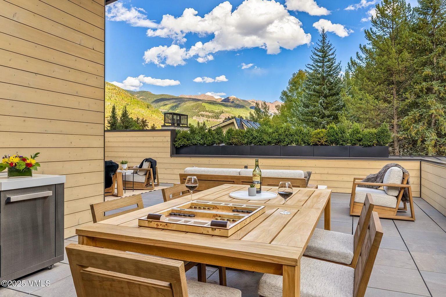 This expansive outdoor deck features a light wood dining set complete with a backgammon board, surrounded by comfortable lounge seating with plush cushions. The space is framed by a low wooden wall and a manicured hedge, offering unobstructed, panoramic views of the surrounding mountains and pine forest. The bright, sunny atmosphere and modern, clean-lined furniture create an inviting setting for al fresco dining and relaxation.