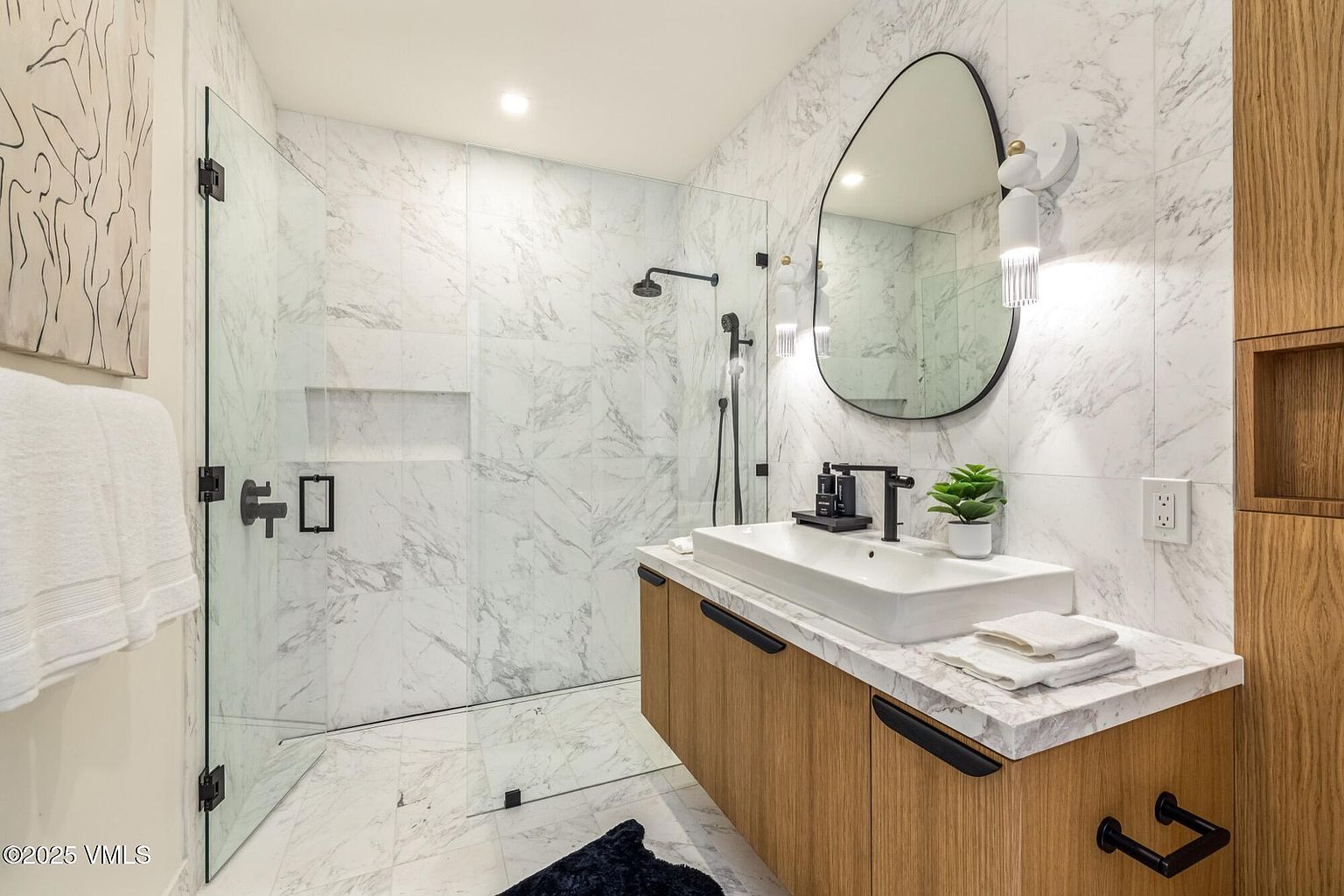This modern bathroom features a sleek, wall-mounted light wood vanity topped with a rectangular vessel sink and a stone countertop. A glass-enclosed walk-in shower with patterned stone wall tiles and a rainfall showerhead sits adjacent to the vanity, which is accented by a uniquely shaped mirror and contemporary lighting. The space is finished with clean lines and a minimalist aesthetic, creating a bright and sophisticated atmosphere.