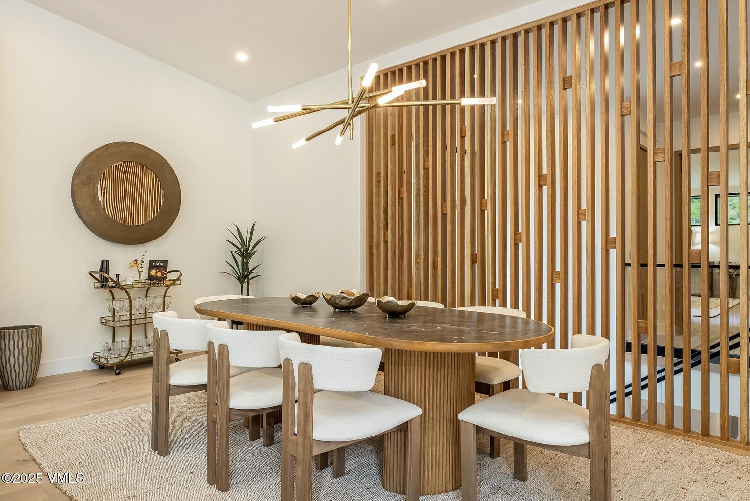 This elegant dining room features a modern oval table with a dark stone top and a fluted base, surrounded by six light-toned chairs with upholstered seats. A striking geometric chandelier hangs above the table, while a decorative wood slat screen provides a semi-private partition to the adjacent space. The room is accented by a circular wall art piece and a gold-toned bar cart, creating a sophisticated and contemporary atmosphere.