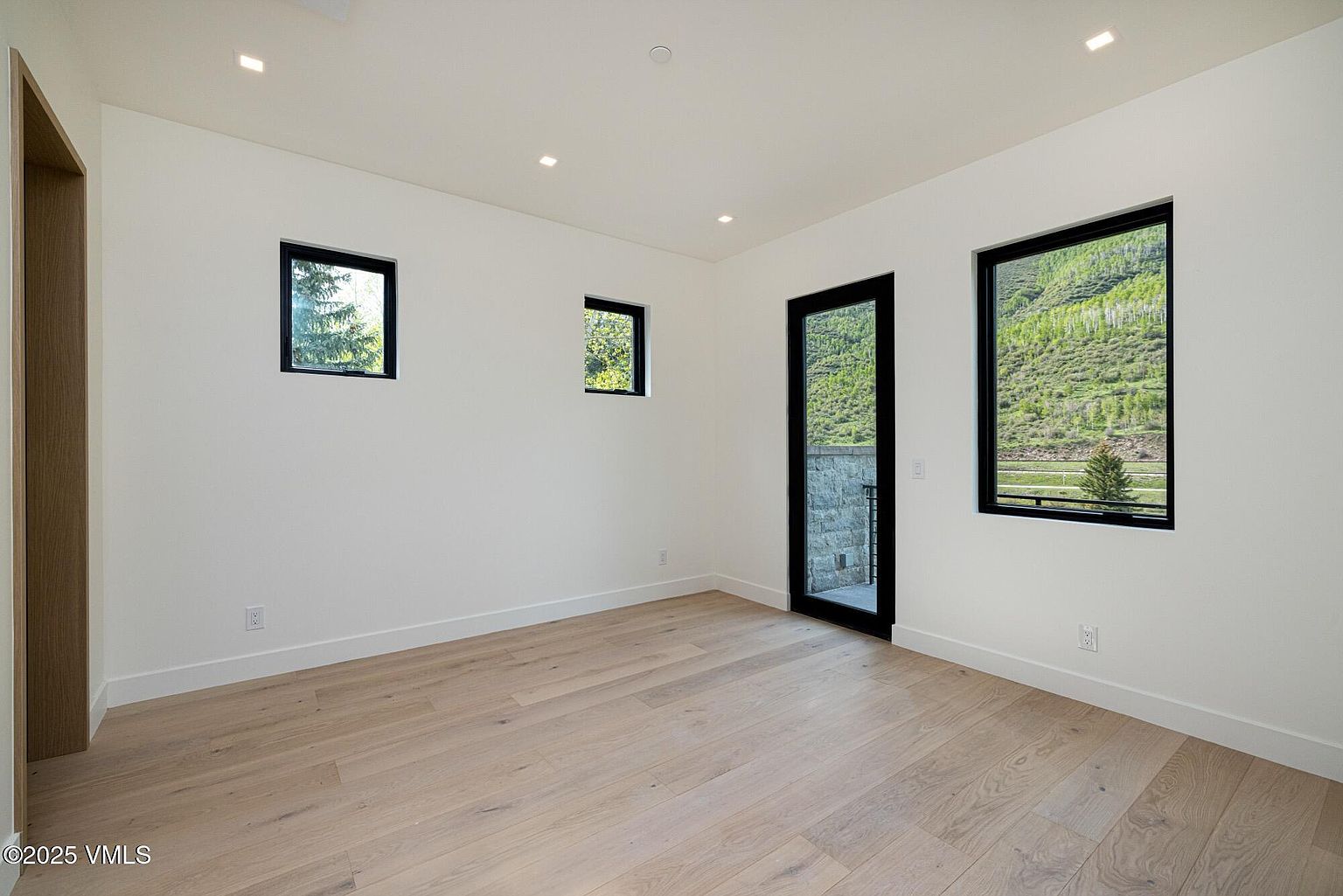 This minimalist, light-filled room features neutral white walls and light wood flooring, creating a clean and airy atmosphere. The space is accented by black-framed windows and a matching glass door that provides a view of the surrounding mountain landscape. Recessed lighting is integrated into the ceiling, enhancing the modern, uncluttered aesthetic of the room.