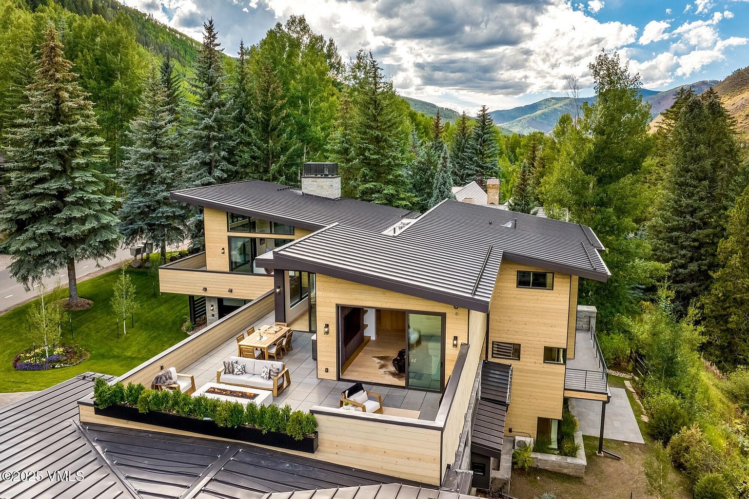 This aerial perspective captures a modern mountain residence characterized by clean lines, light wood siding, and a dark metal roof. The home features a spacious, multi-level terrace equipped with an outdoor dining area, a lounge space with a fire pit, and integrated planters. Surrounded by lush evergreen trees and set against a backdrop of rolling mountains, the property offers a seamless blend of contemporary architecture and natural beauty.