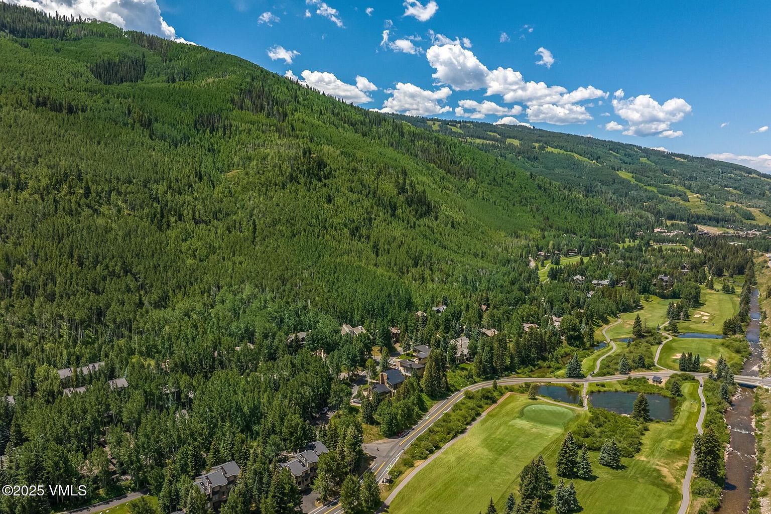 This high-angle aerial view captures a scenic mountain landscape featuring a lush, forested hillside rising above a residential community. In the foreground, a manicured golf course with a small pond and a winding road integrates seamlessly into the natural environment. The perspective highlights the expansive scale of the property's surroundings, emphasizing the peaceful, nature-focused setting.