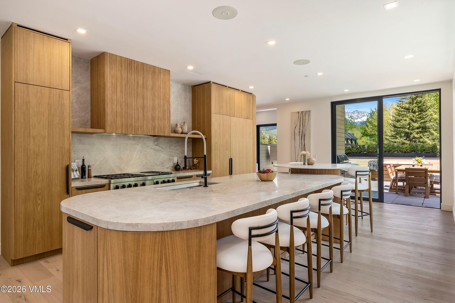 This modern kitchen features light wood cabinetry and a large, curved island topped with a light-toned stone surface, providing ample seating with four bar stools. A professional-style range is set against a stone backsplash, while large glass doors open to an outdoor deck, blending indoor and outdoor living spaces. The room is illuminated by recessed lighting and offers a bright, airy atmosphere with a view of the surrounding landscape.