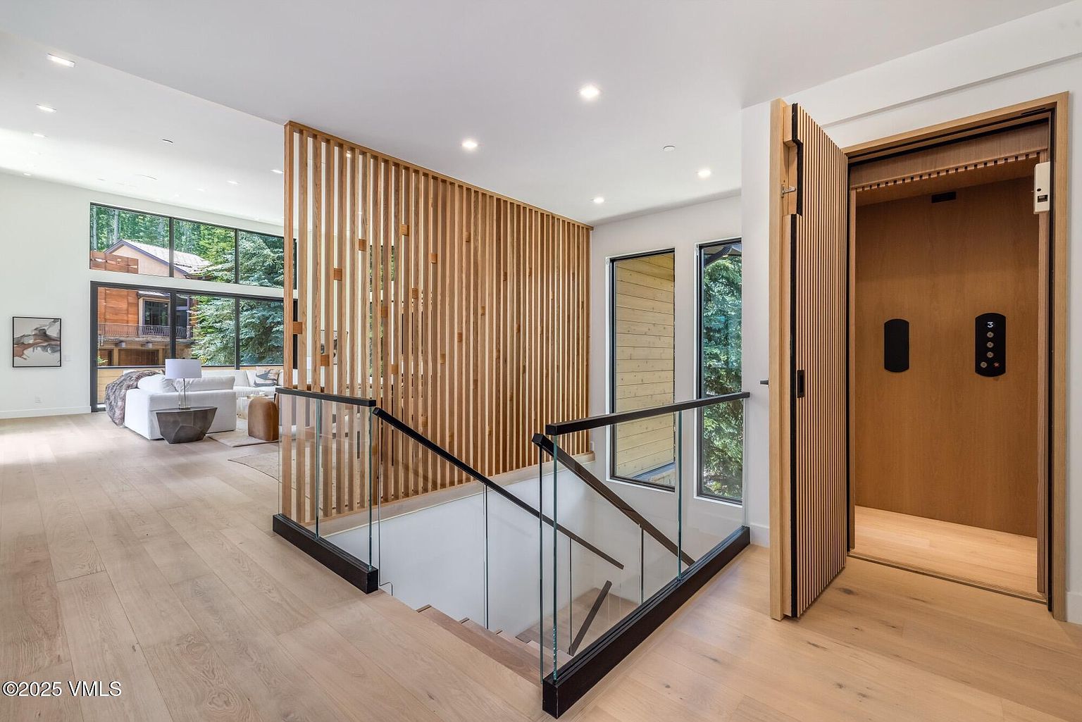 This modern interior space features a staircase with glass railings and a striking vertical wood slat partition that separates the landing from the living area. To the right, a wood-paneled elevator provides a sleek, functional design element, while light wood flooring runs throughout the open-concept layout. Large windows allow natural light to fill the space, offering a view of the surrounding trees and neighboring architecture.