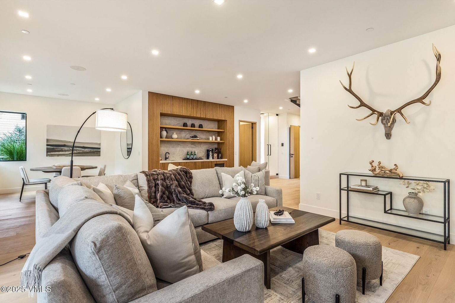 This bright and inviting living room features a large, plush sectional sofa adorned with a textured throw blanket and decorative pillows. A dark wood coffee table sits atop a neutral area rug, flanked by two upholstered ottomans, while a sleek metal console table displays decorative objects beneath a prominent antler wall mount. In the background, a custom wood-slatted wall unit with built-in shelving adds warmth and architectural interest to the open-concept space.