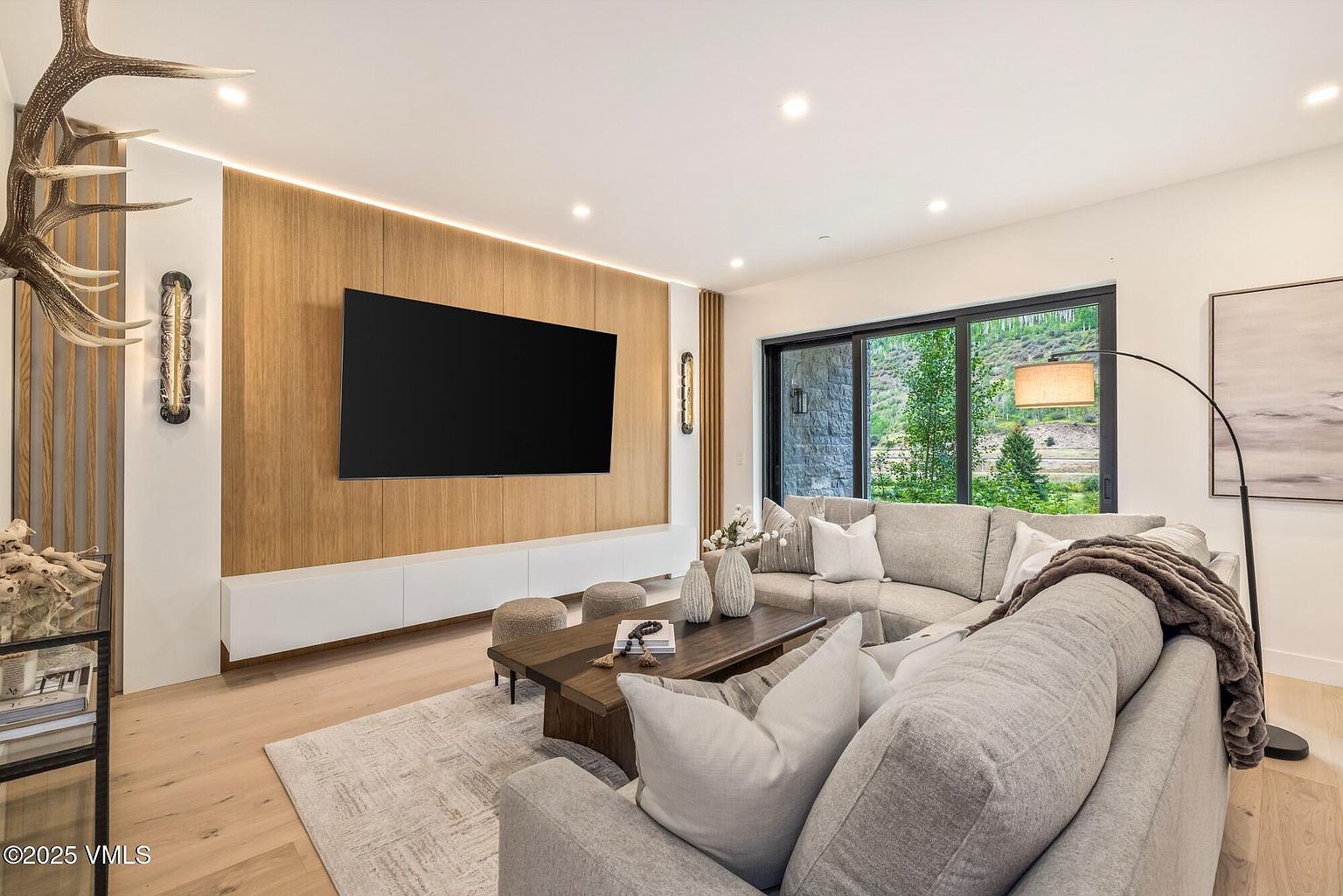 This contemporary living room features a large, light-toned sectional sofa arranged around a dark wood coffee table on a textured area rug. The focal point is a wall with vertical wood paneling and integrated lighting, housing a large flat-screen television above a sleek, white low-profile media console. Large sliding glass doors provide a view of the natural landscape outside, while a tall floor lamp and decorative antler sculpture add unique character to the space.
