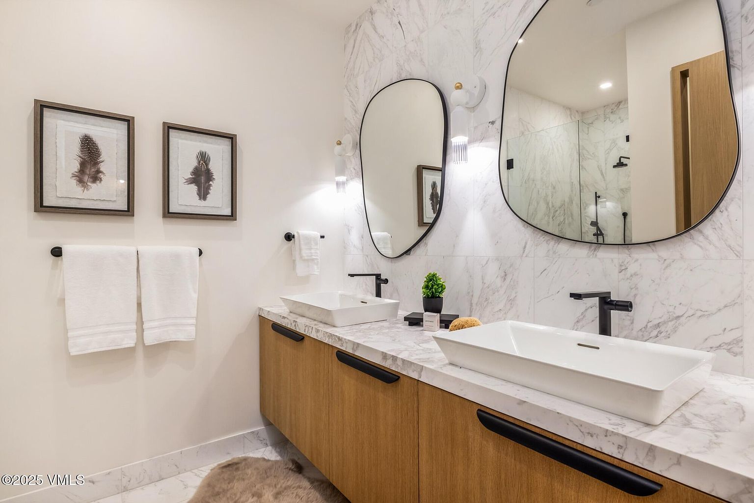This modern primary bathroom features a floating vanity with light wood cabinetry and a stone countertop, complemented by two rectangular vessel sinks and matte black fixtures. The wall is adorned with large-format stone tiles, accented by two uniquely shaped, asymmetrical mirrors and minimalist wall sconces. The space is finished with framed wall art and a soft rug, creating a sophisticated and contemporary aesthetic.