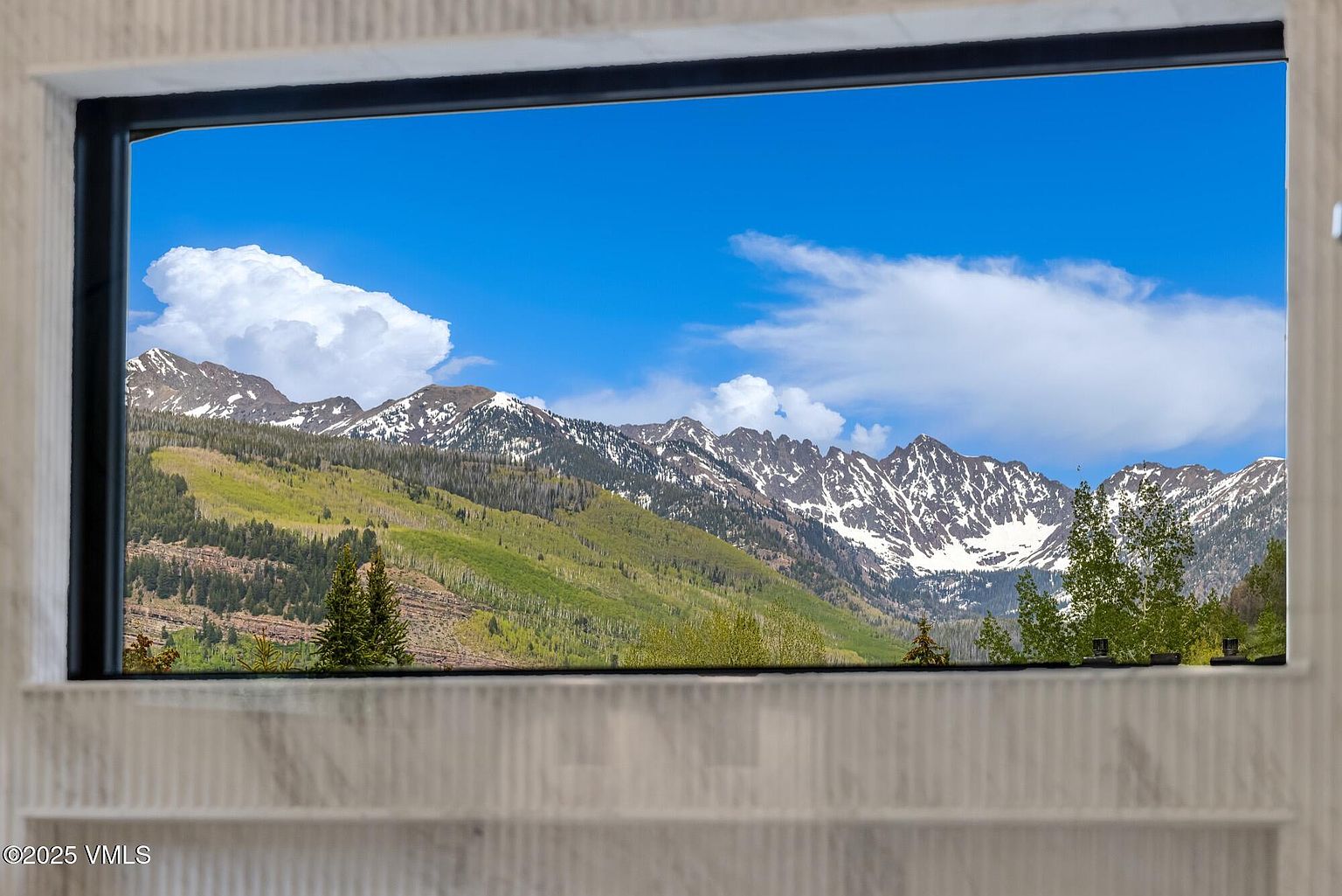 This image captures a large, rectangular window framed by a light-toned, textured stone surround that serves as a focal point. Through the glass, a breathtaking panoramic view of rugged, snow-dusted mountain peaks and lush green slopes is showcased, creating a natural piece of art. The perspective is centered and cinematic, emphasizing the seamless integration of the interior architecture with the dramatic outdoor landscape.