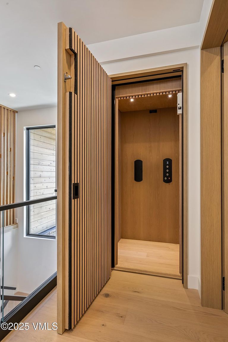 This interior view highlights a modern home elevator accessed through a sleek, slatted wood-paneled door that swings open to reveal a warm, wood-lined interior. To the left, a glass-railed staircase descends, offering a glimpse of a window with horizontal wood siding visible outside. The aesthetic is minimalist and contemporary, emphasizing clean lines and high-quality natural wood finishes.