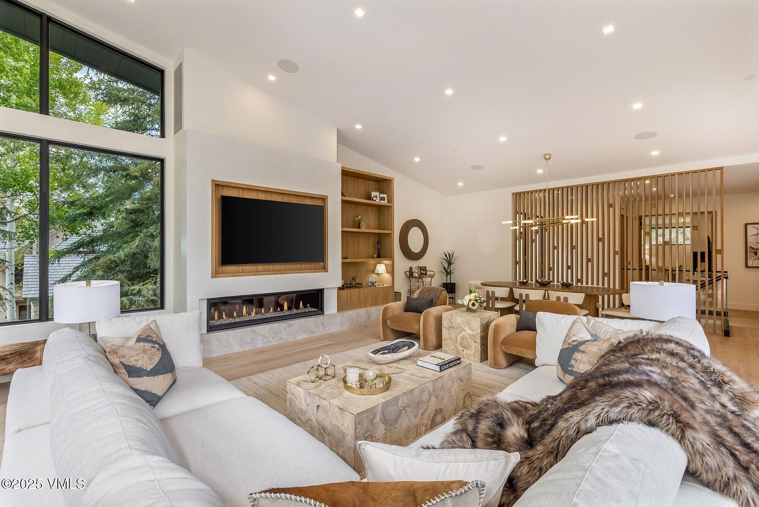 This bright and airy living room features a contemporary design with a large, light-colored sectional sofa and a unique, textured wood coffee table. A linear fireplace is set into a stone-clad wall, which also incorporates built-in shelving and a mounted television. Large windows provide ample natural light and views of the surrounding trees, while a dining area with a slatted wood partition is visible in the background.