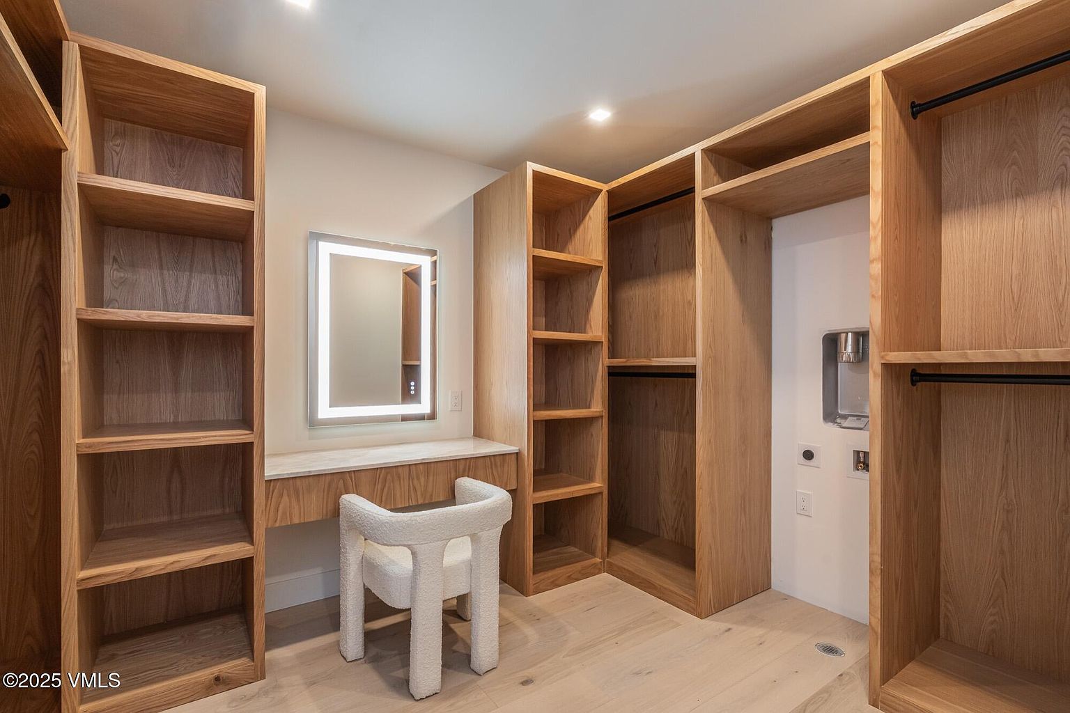 This walk-in closet features custom light wood cabinetry with extensive shelving and hanging space, providing ample storage organization. A built-in vanity area with a backlit mirror and a textured white chair creates a functional dressing space. The room is finished with light-toned wood flooring and recessed lighting, offering a clean and modern aesthetic.
