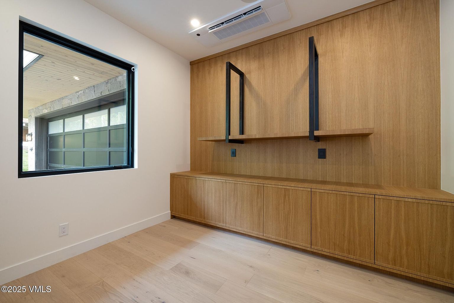 This modern office space features light wood flooring and a wall-to-wall built-in storage unit crafted from light wood cabinetry. The wall includes integrated shelving supported by black metal brackets, providing a clean and contemporary aesthetic. A large window to the left offers a view of an exterior garage area, filling the room with natural light.