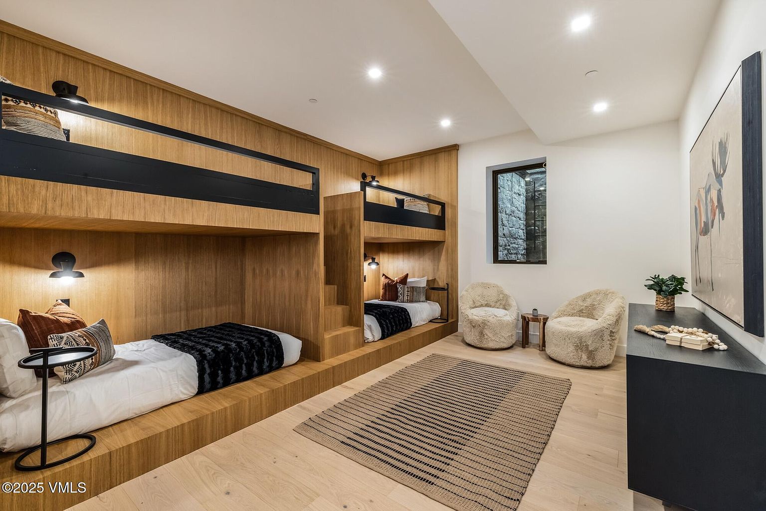 This modern bedroom features custom built-in bunk beds crafted from light wood, providing a cozy and efficient sleeping arrangement. The room includes two plush, textured armchairs positioned near a window, a striped area rug, and a sleek black console table adorned with decorative items. The clean lines and warm wood tones create a sophisticated yet inviting atmosphere suitable for a guest suite or children's room.