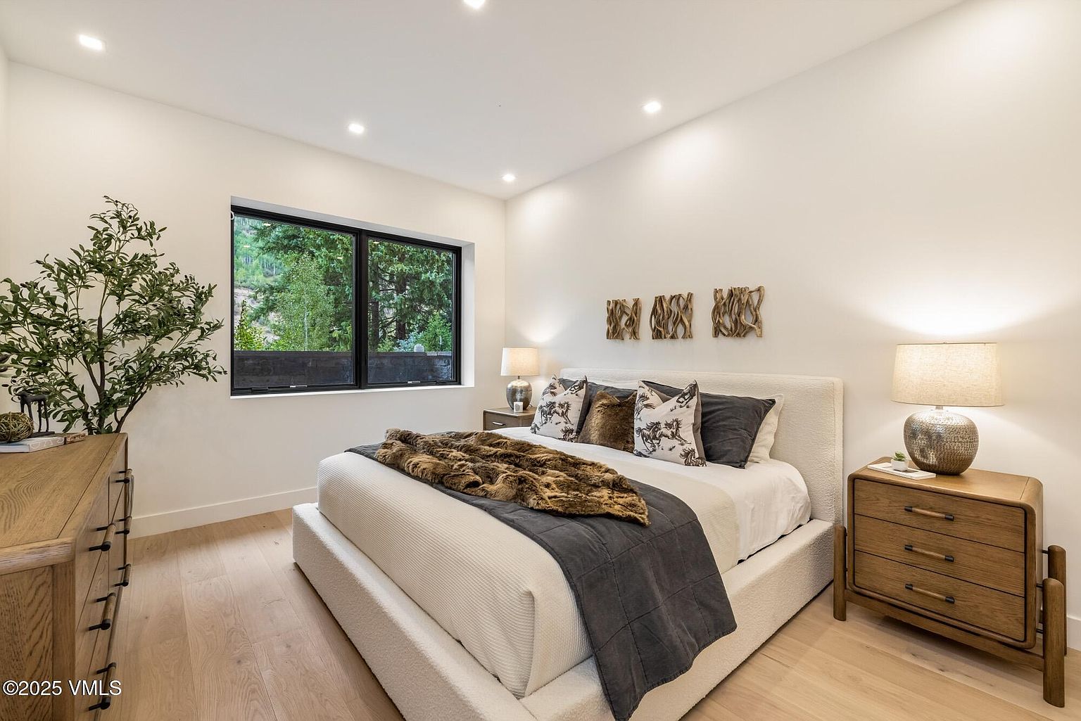 This modern primary bedroom features a plush, light-colored upholstered bed frame adorned with a textured faux-fur throw and patterned accent pillows. The room is anchored by light wood flooring and a matching wooden nightstand, while a large window provides a view of the surrounding trees. Three decorative wooden wall art pieces hang above the headboard, contributing to the serene and contemporary aesthetic.