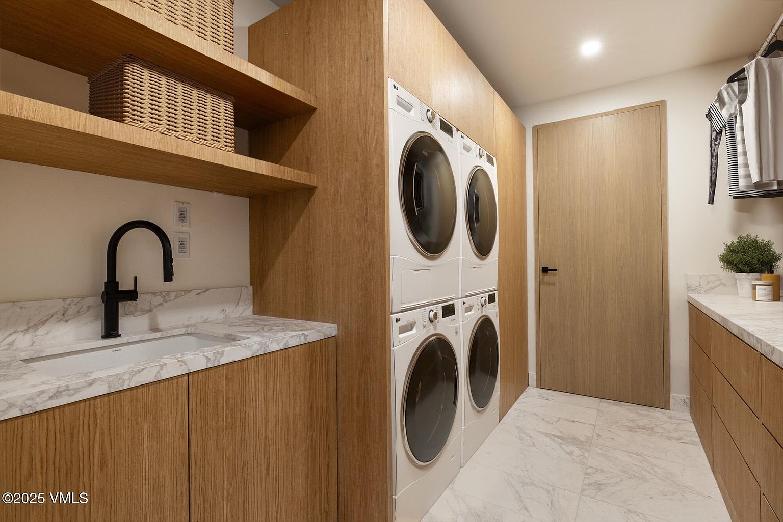 This modern laundry room features light wood cabinetry and a stone countertop with an integrated sink and a sleek black faucet. The space is equipped with a stacked washer and dryer set, providing a clean and efficient layout. The room is finished with light-toned stone flooring and open shelving, creating a bright and organized utility area.