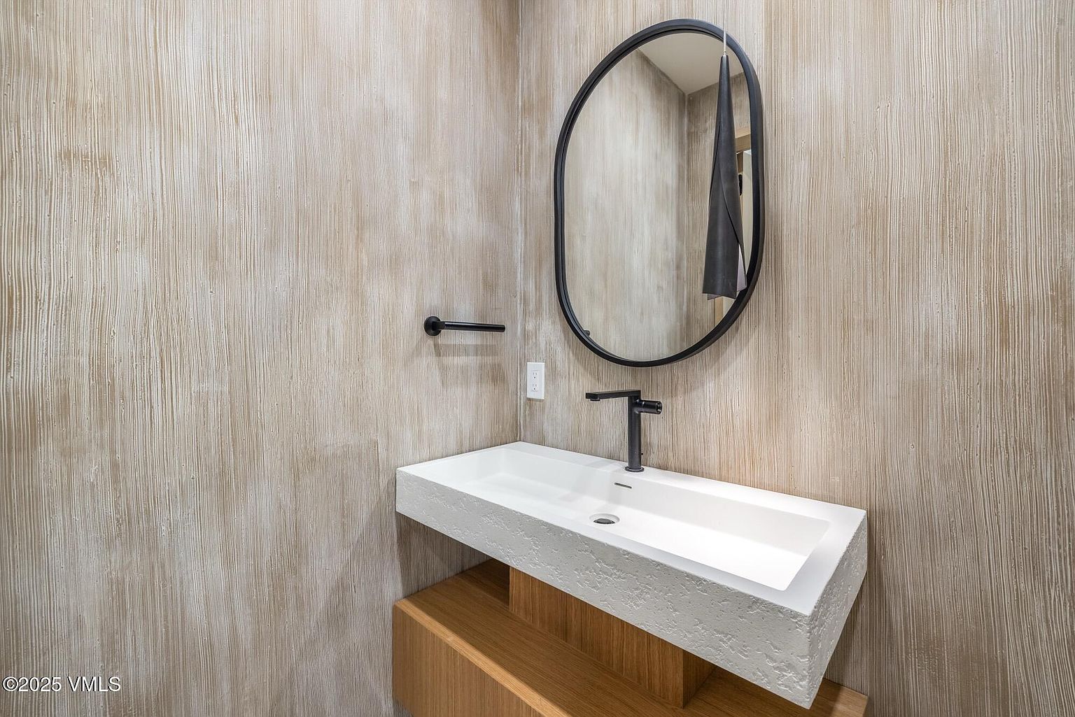 This modern powder room features a unique, textured wall treatment that adds depth and visual interest to the space. A minimalist, rectangular white sink with a rough-hewn edge sits atop a light wood vanity, complemented by a sleek black faucet and a matching oval mirror. The overall aesthetic is contemporary and sophisticated, emphasizing clean lines and a neutral, earthy color palette.