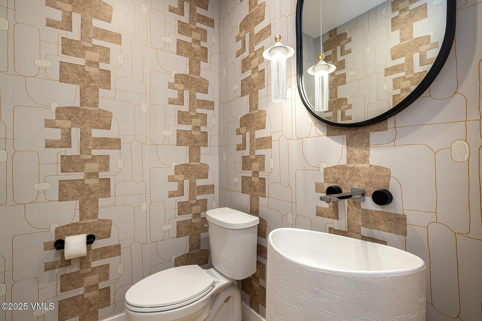 This powder room features a striking, textured wallpaper with a geometric pattern in neutral earth tones. A unique, cylindrical pedestal sink stands beside a modern toilet, complemented by a wall-mounted faucet and a sleek, oval-shaped mirror. Two elegant, tiered pendant lights hang above the vanity, creating a sophisticated and artistic atmosphere.