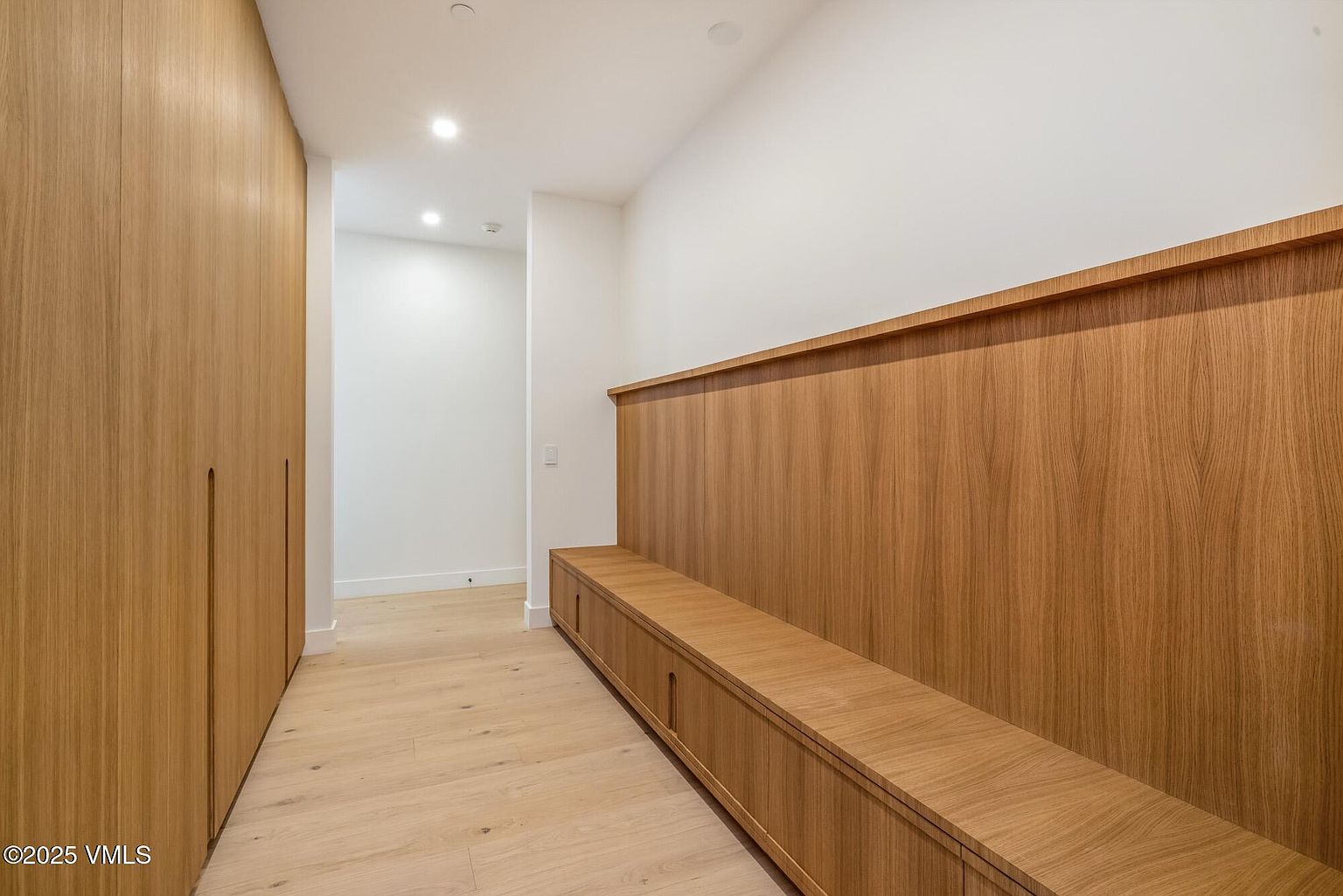 This minimalist hallway features light wood flooring and extensive built-in cabinetry with a matching wood finish. A long, integrated bench with storage cabinets runs along the right wall, topped by a continuous wood paneling detail that creates a clean, cohesive aesthetic. The space is illuminated by recessed ceiling lights, leading toward a bright, neutral-toned transition area.
