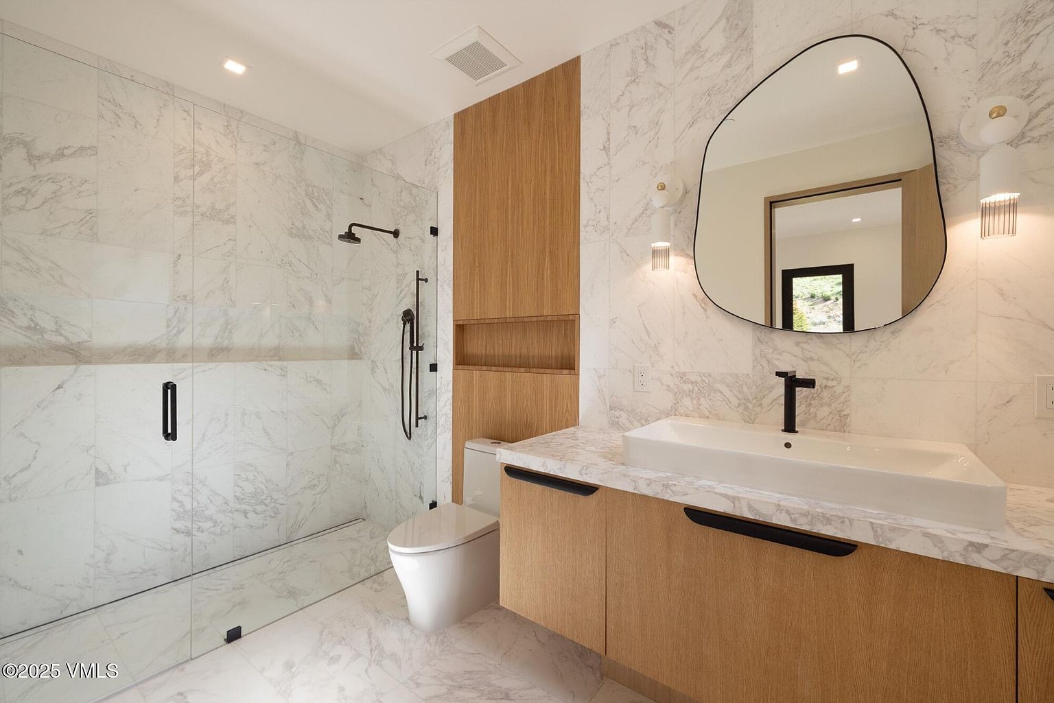 This modern bathroom features a sleek vanity with light wood cabinetry and a stone countertop, paired with a unique, organic-shaped mirror. To the left, a glass-enclosed shower is finished with light-toned stone tile, while a wall-mounted toilet sits adjacent to a recessed wood-lined storage niche. The space is illuminated by contemporary wall sconces, creating a clean and sophisticated aesthetic.
