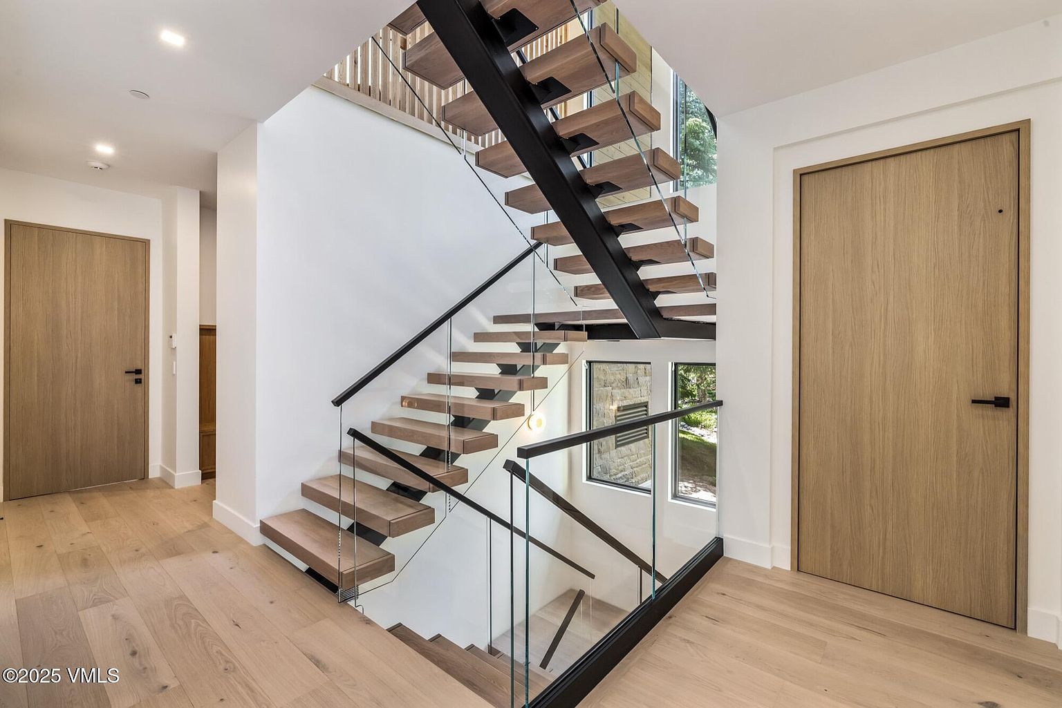 This modern interior features a striking floating staircase with light wood treads and a black metal frame, complemented by glass railings that maintain an open, airy feel. The space is finished with light wood flooring and crisp white walls, creating a clean, contemporary aesthetic. Large windows at the landing allow natural light to filter through, highlighting the architectural design of the staircase.