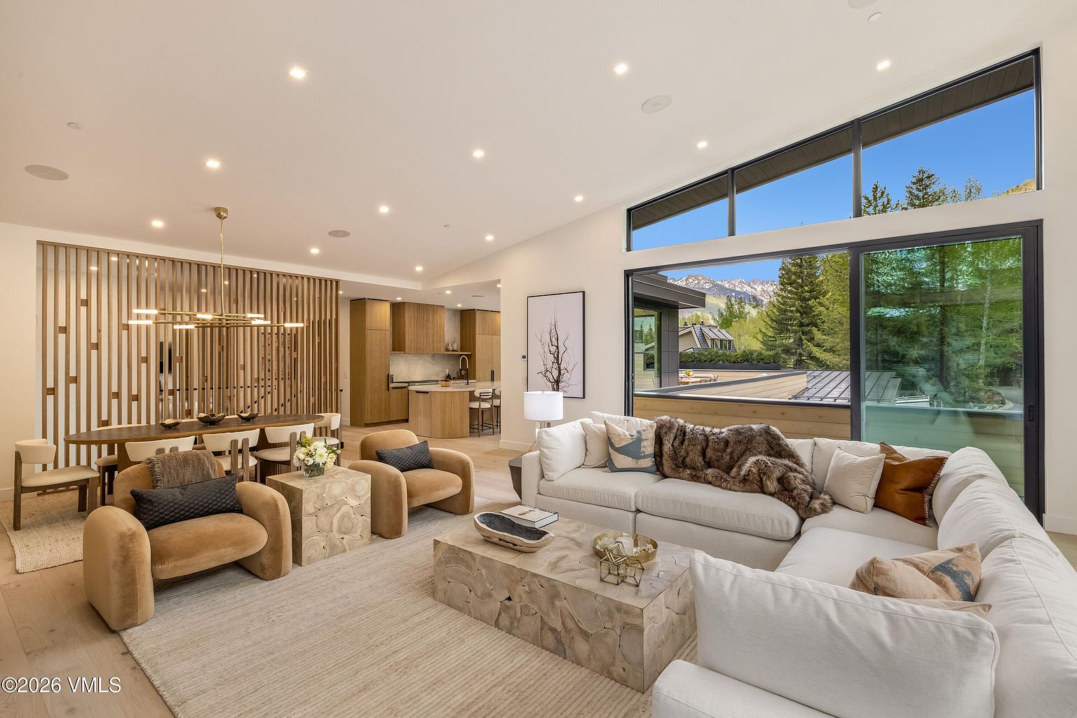 This open-concept living area features a contemporary design with a large white sectional sofa, two plush tan armchairs, and a unique carved wood coffee table. The space is anchored by a light-toned area rug and flows seamlessly into a dining area defined by a decorative wood-slat partition and a modern chandelier. Large windows provide an expansive view of the surrounding trees and distant mountains, creating a bright and airy atmosphere.