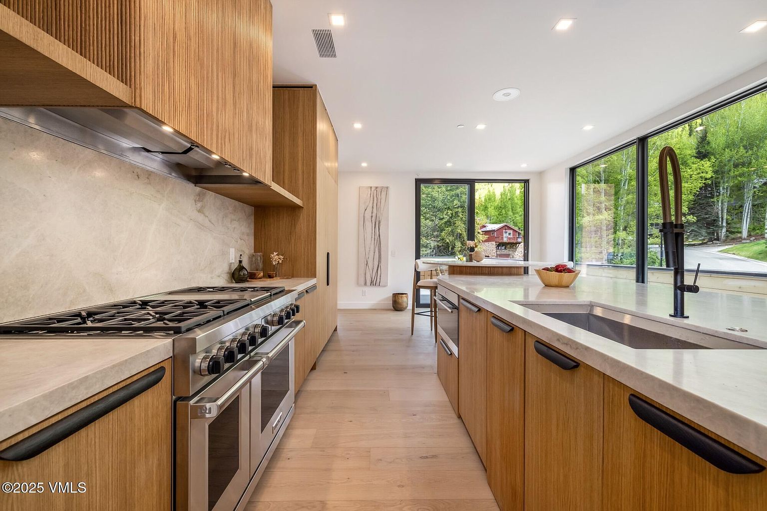This modern kitchen features sleek light wood cabinetry, a large central island with a stone countertop, and high-end stainless steel appliances. A long, expansive window provides a bright, scenic view of the surrounding forest, while the minimalist design creates a clean and sophisticated atmosphere. The perspective looks directly down the galley-style layout, highlighting the seamless integration of the cooking area and the adjacent dining space.