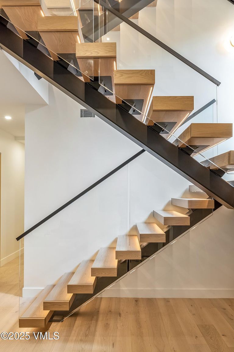 This image captures a modern, floating staircase featuring light wood treads and a sleek black metal stringer. A glass railing system provides an open, airy feel, while integrated lighting under each step adds a sophisticated, warm glow. The perspective is from the base of the stairs, looking upward to highlight the architectural geometry and clean lines of the design.