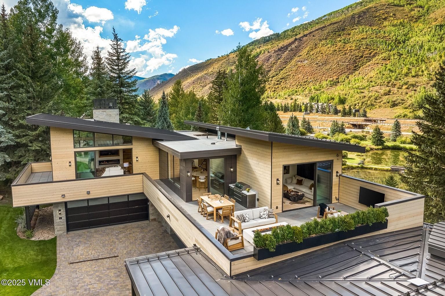 This aerial perspective captures a contemporary mountain residence featuring a multi-level design with light wood siding and dark metal roofing. The home includes a spacious elevated deck furnished with outdoor seating and a dining area, overlooking a scenic landscape with trees and a pond. The architectural style emphasizes clean lines and large glass openings that seamlessly connect the indoor living spaces to the expansive outdoor terrace.