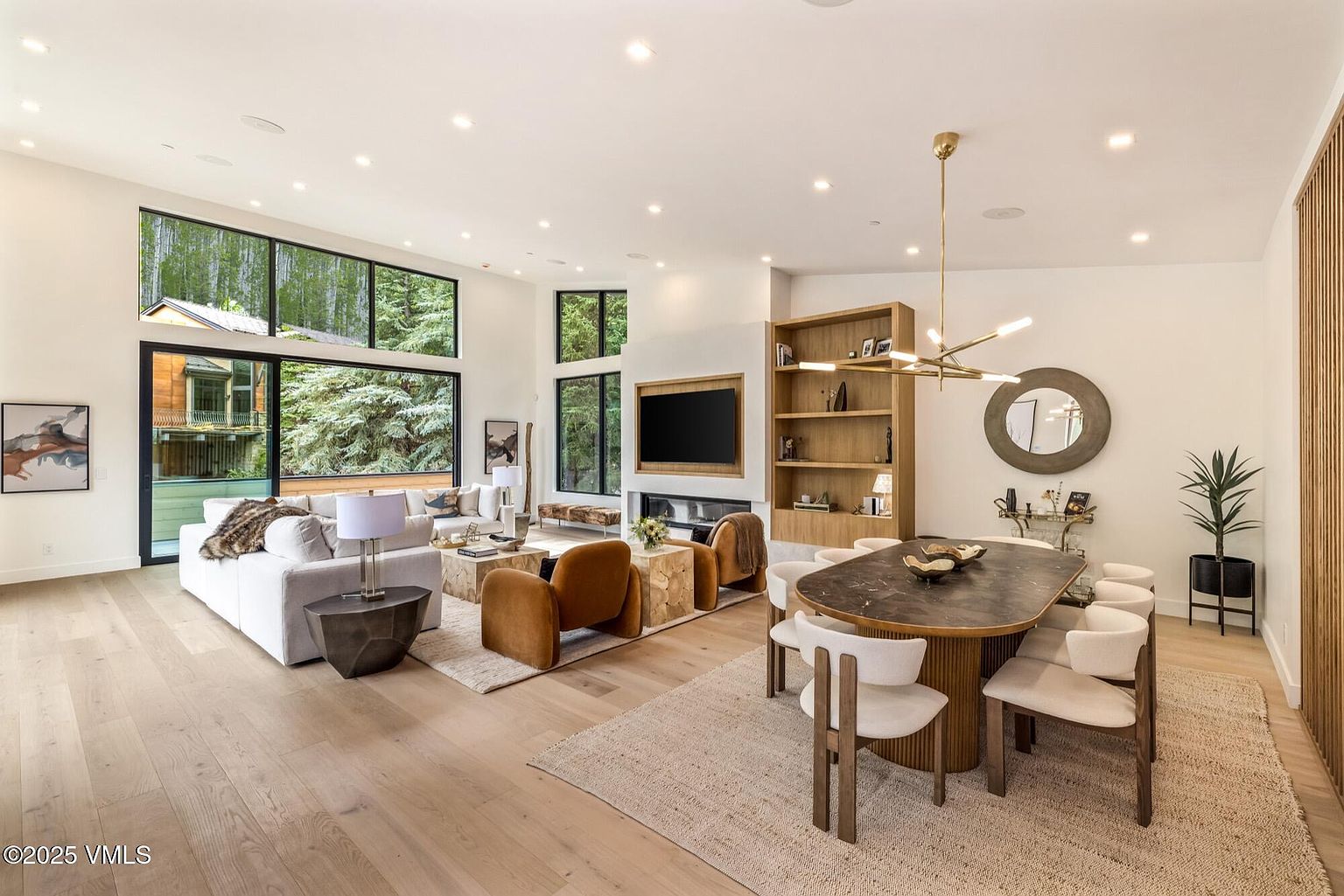 This open-concept living and dining area features light wood flooring and a bright, airy atmosphere enhanced by large windows that frame the surrounding forest. The living space is anchored by a central fireplace with a television mounted above, flanked by a tall wooden shelving unit, while the dining area showcases an oval table with modern chairs beneath a sculptural gold chandelier. The room is decorated in a sophisticated, neutral palette with warm textures, creating a cohesive and inviting space for entertaining.