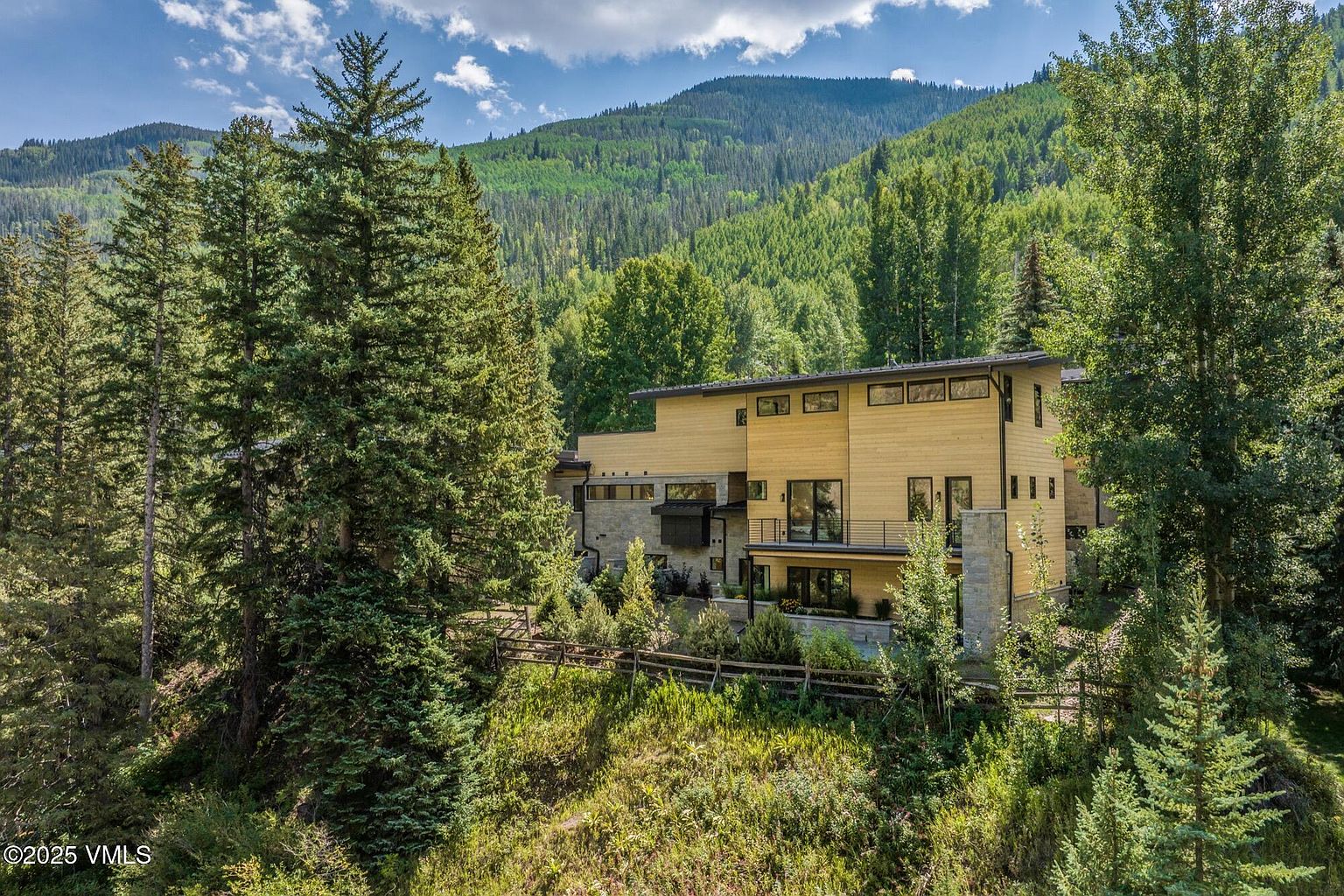 This elevated perspective captures a modern mountain residence nestled within a dense forest of pine and aspen trees. The home features a multi-level design with a combination of horizontal wood siding and stone accents, complemented by a balcony and large windows that overlook the surrounding landscape. The setting is serene and secluded, emphasizing the home's integration into the natural, mountainous terrain.