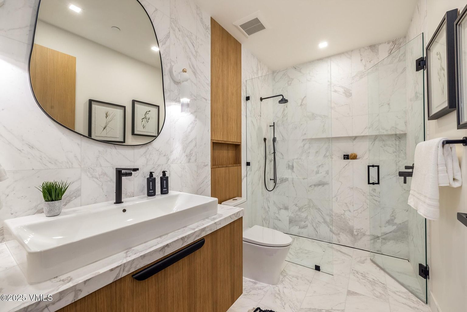 This modern bathroom features a sleek vanity with a light wood finish and a white rectangular sink, complemented by a large, uniquely shaped mirror. The space is finished with light-toned stone wall and floor tiles, and includes a glass-enclosed walk-in shower with matte black fixtures. A built-in wood-toned storage niche adds warmth and functionality to the contemporary design.