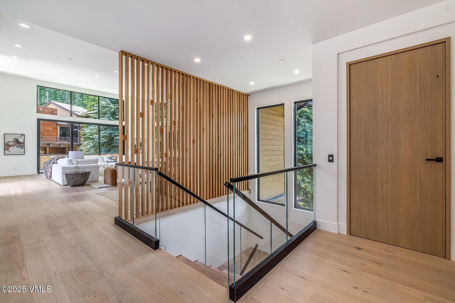 This modern entryway features a striking vertical wood slat screen that separates the staircase from the adjacent living area. The space is characterized by light wood flooring, clean white walls, and a glass-railed staircase that leads to a lower level. Large windows provide ample natural light and a view of the wooded exterior, creating an open and airy atmosphere.