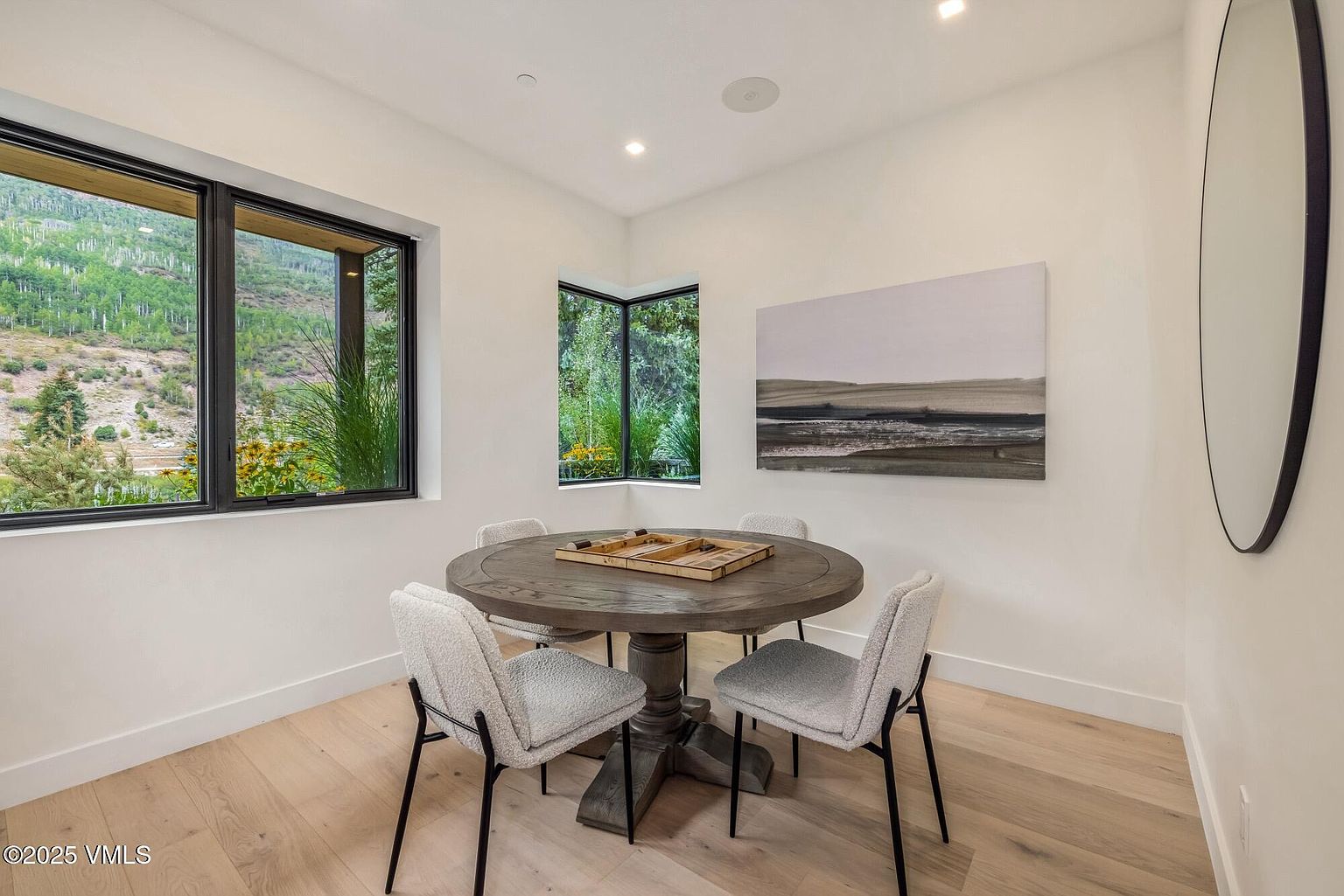 This bright dining area features a round wooden table centered in the room, surrounded by four modern chairs with light-colored textured upholstery and dark metal legs. A large window and a corner window provide expansive views of the lush, mountainous landscape outside, filling the space with natural light. The room is finished with light wood flooring, white walls, and a large abstract piece of wall art, creating a serene and minimalist atmosphere.