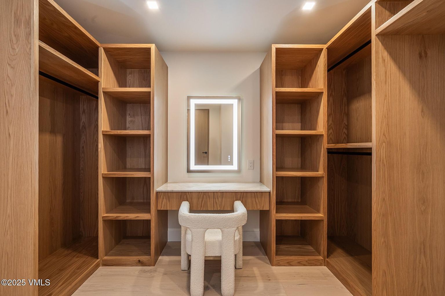 This walk-in closet features custom light wood cabinetry with open shelving and hanging space on both sides. A central vanity area includes a stone-topped desk, a modern illuminated mirror, and a textured white chair, creating a functional and elegant dressing space. The room is brightly lit by recessed ceiling lights, emphasizing the clean lines and organized layout.
