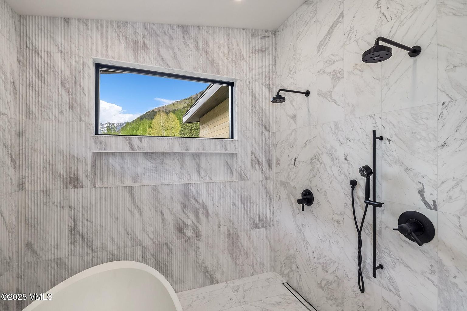 This modern bathroom features a walk-in shower area with light-colored, veined stone wall tiles and a recessed niche for storage. The shower is equipped with matte black fixtures, including a rainfall showerhead, a handheld sprayer, and a wall-mounted control. A rectangular window provides a scenic view of the mountains, while a portion of a white soaking tub is visible in the foreground.