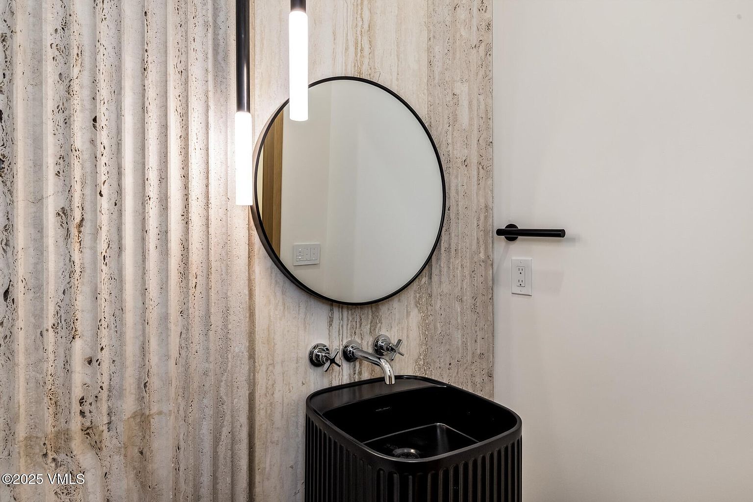 This modern powder room features a striking textured stone wall with vertical fluting that serves as a dramatic backdrop for a circular mirror. A sleek, black, vertically-ribbed vanity cabinet supports a wall-mounted chrome faucet, while minimalist pendant lights hang on the left. The space is finished with a clean white wall on the right, creating a sophisticated and high-contrast aesthetic.