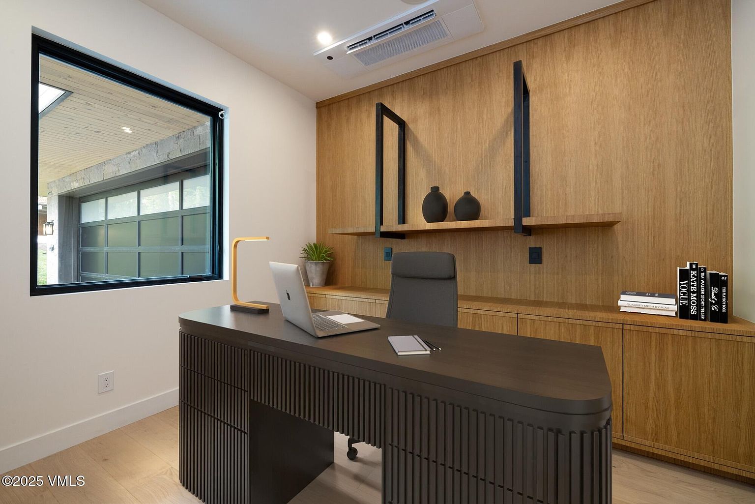 This contemporary home office features a sleek, dark-toned desk with a textured, slatted base positioned centrally in the room. A wall of light wood paneling serves as a backdrop, incorporating built-in cabinetry and open shelving accented by black metal supports. A large window to the left provides natural light and a view of an adjacent structure, while the minimalist decor and clean lines create a professional and sophisticated workspace.