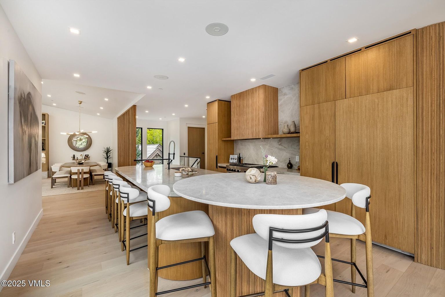 This modern, open-concept kitchen features a large, circular-ended island with a light stone countertop and light wood cabinetry. The space is accented by vertical wood paneling on the cabinetry and island base, creating a warm, textured aesthetic. In the background, a dining area is visible, and the room is illuminated by recessed lighting and a contemporary chandelier.