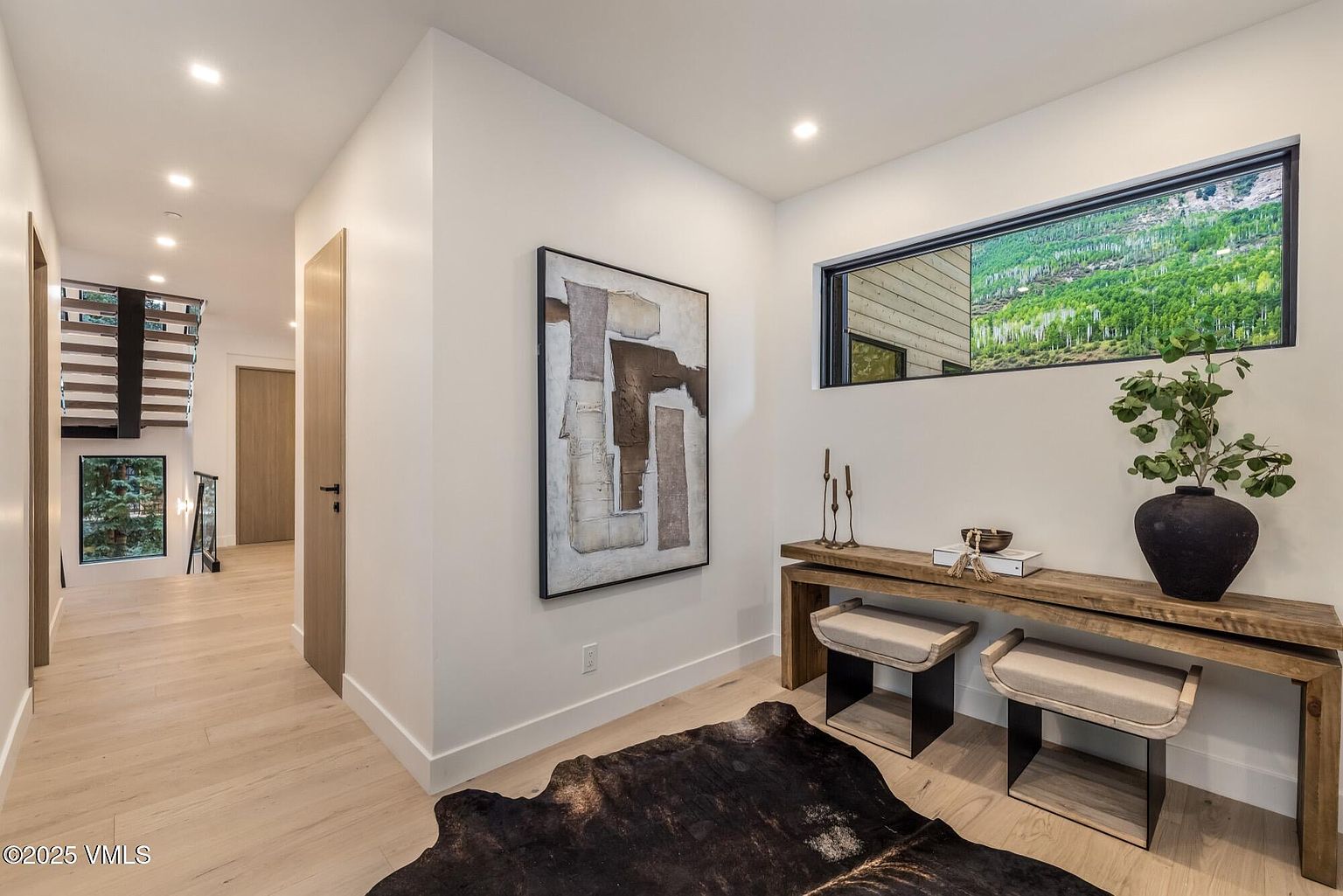 This elegant entryway features light wood flooring and a neutral color palette, creating a bright and welcoming atmosphere. A rustic wooden console table sits beneath a horizontal window that frames a scenic outdoor view, complemented by a large abstract piece of wall art. The space flows into a hallway that leads to a staircase, showcasing a modern design with clean lines and natural textures.